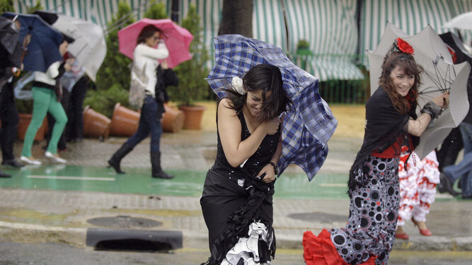 Dos mujeres vestidas de flamenca se intentan refugiar de la lluvia y el viento bajo un paraguas.