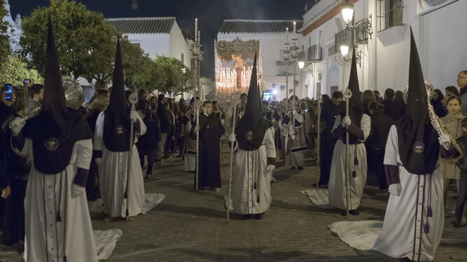 La cofradía de la Soledad de Olivares cruza la Plaza de España de este municipio aljarafeño. Los nazarenos arrastran la cola de sus túnicas. Al fondo, el elegante palio de la Virgen de los Dolores.