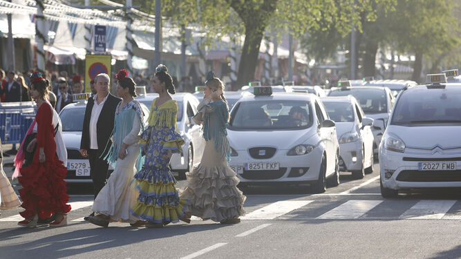 Varias mujeres vestidas de flamenca cruzan el paso de peatones junto a la parada de taxis de la Feria.