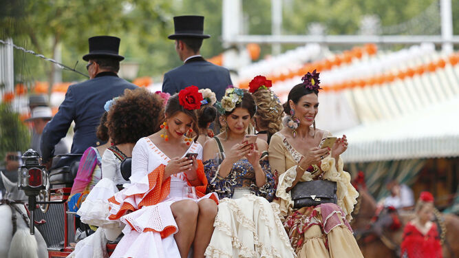 Mujeres de flamenca consultan el móvil mientras pasean en coche de caballo.