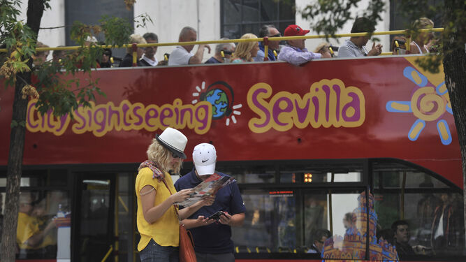 Turistas mirando el mapa de la ciudad hispalense y detrás un autobús turístico