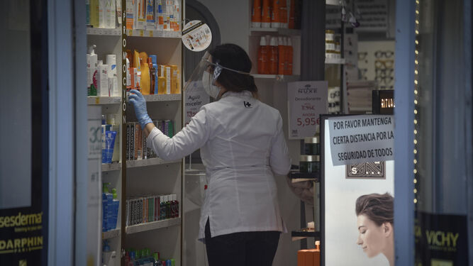 Una farmacéutica provista de mascarilla y pantalla protectora coloca productos en una estantería.