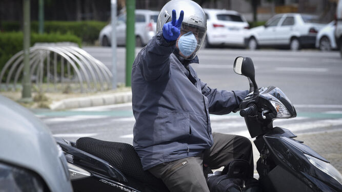 Un hombre equipado con mascarilla y guantes hace con su mano desde su moto el signo de la victoria.