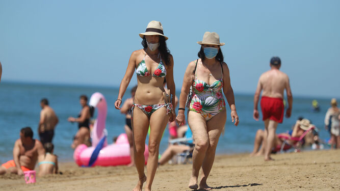 Dos bañistas pasean por la playa onubense de el Portil con sombrero y mascarilla.