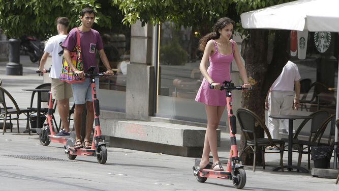Dos usuarios en sendos patinetes de alquiler por el carril bici de la Avenida de la Constitución de Sevilla, ambos sin casco protector.