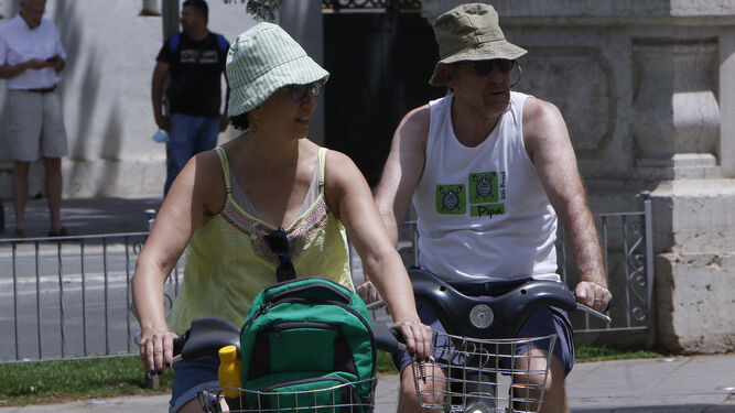 Dos turistas con gorro montados en bici al sol.