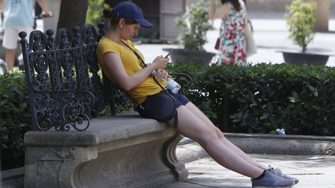 Una joven mira el móvil en un banco a la sombra.