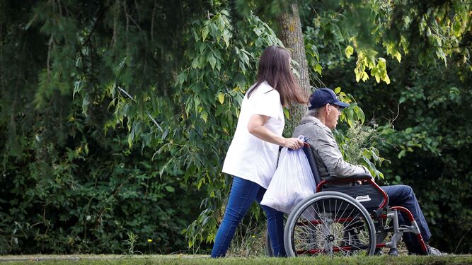 Una mujer pasea con un hombre en silla de ruedas junto a una residencia  de mayores.