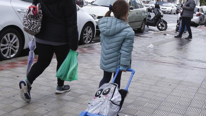 Una menor junto a su madre a la salida tira del carrito con su mochila a la salida del colegio.
